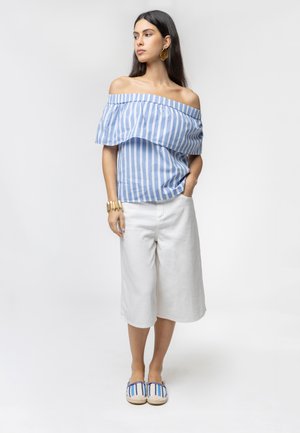 Donna che indossa un top rigato blu e bianco a spalle scoperte, culotte bianchi, scarpe slip-on rigate, braccialetto d'oro e grandi orecchini d'oro.