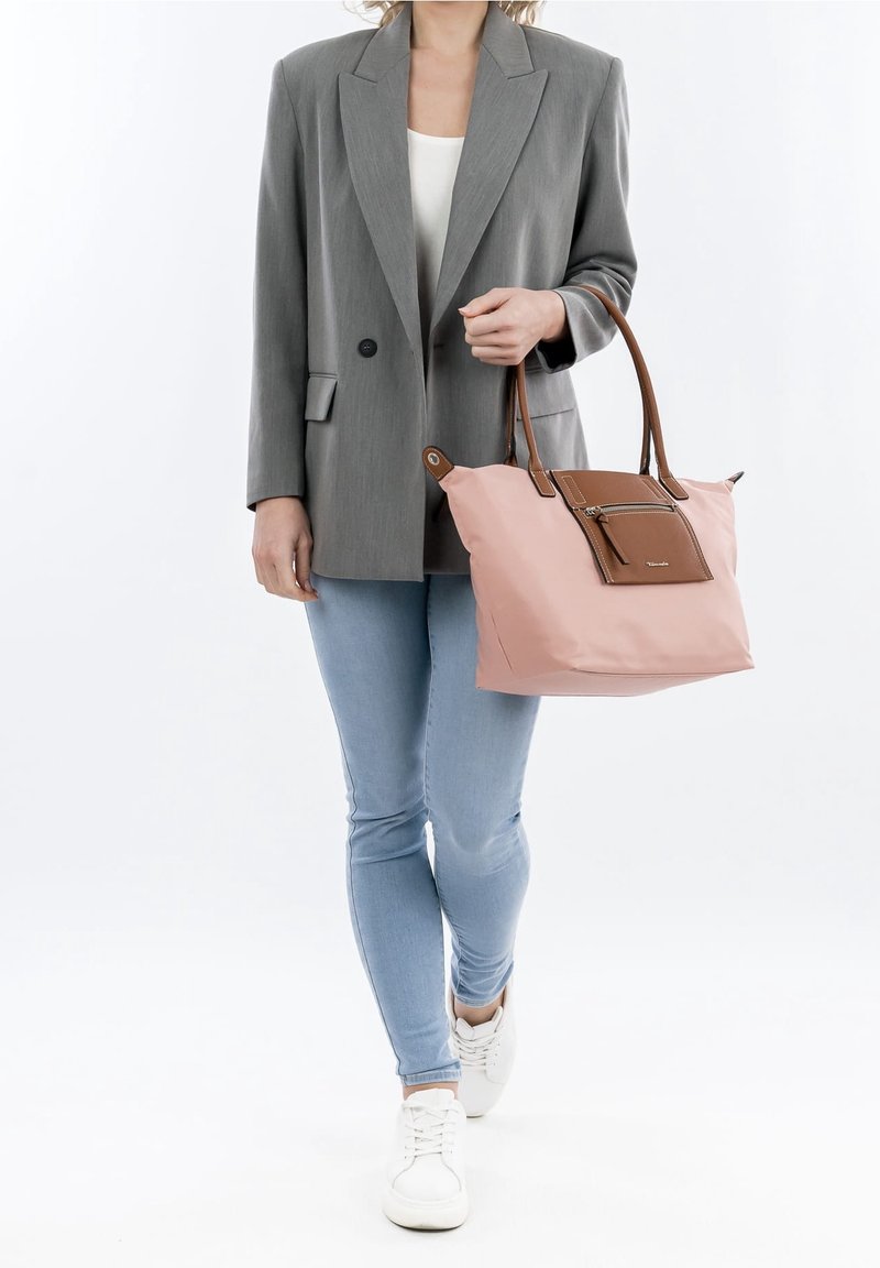 Femme portant un blazer gris, une chemise blanche, un jean bleu clair et des baskets blanches, tenant à la main un sac à main rose et marron.
