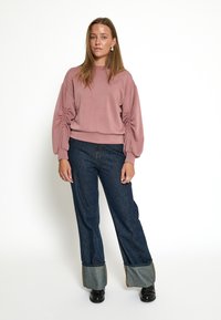 Svetloružový oversized sweatshirt s naťahovacími rukávmi, spárovaný so tmavomodrými rozšírenými džínsami s kontrastnými denimovými manžetami a čiernymi topánkami.