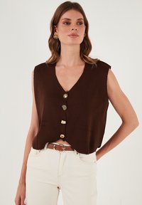 Brązowa bezrękawnik z dekoltem w kształcie litery V, z pięcioma ozdobnymi guzikami i dwiema przednimi kieszeniami. Połączona z jasnobeżowymi dżinsami i paskiem.