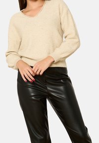 Beige gestrickter Pullover mit V-Ausschnitt und langen Ärmeln, der eine strukturierte Oberfläche aufweist. Kombiniert mit schwarzen Kunstlederhosen.