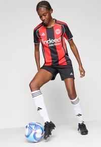 Juodas ir raudonas dryžuotas futbolo marškinėliai su "indeed" logotipu, derinami su juodais šortais ir baltais kojiniais su juodais dryžiais, padėti virš daugiaspalvio kamuolio.