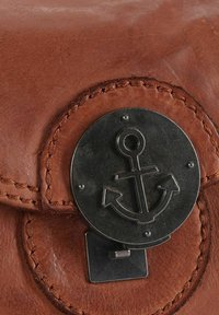 El cierre de la bolsa de cuero marrón presenta una pieza de metal redonda con un diseño de ancla. El detalle de costuras alrededor del borde añade textura.