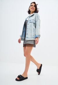 Giacca in denim chiaro con tasche frontali, sopra un vestito in rete nero, abbinata a sandali neri con fascetta decorativa e dettagli in argento.