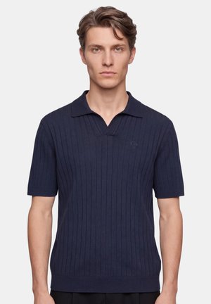 Junger Mann mit kurzen braunen Haaren, der ein dunkelblaues, geripptes Poloshirt mit kurzen Ärmeln trägt und mit dem Gesicht nach vorne vor einem einfarbigen Hintergrund steht.