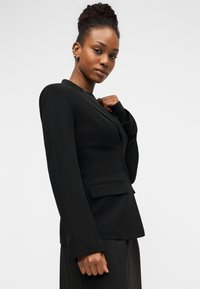 Blazer noir ajusté avec une silhouette cintrée, des épaules structurées et deux poches avant. Tissu lisse à finition mate.