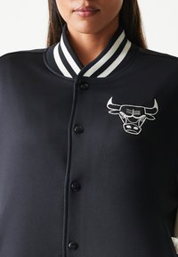 Μαύρο μπουφάν με ribbed μαύρο και λευκό γιακά, με λευκό κεντητό λογότυπο ταύρου στο αριστερό στήθος. Λεία υφή, κλείσιμο με κουμπιά.