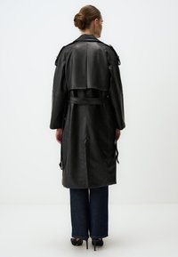 Cappotto in pelle nera con vita cinturata, maniche larghe e retro corto. Abbinato a pantaloni in denim blu e tacchi neri.