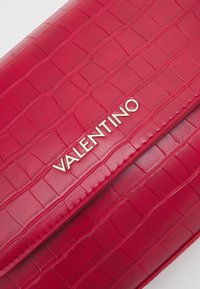 Červená kožená kabelka s texturou krokodýlí kůže, která na přední straně nese zlaté logo "VALENTINO" a má hladký povrch.