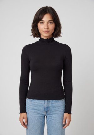 Vrouw met schouderlang donker haar, die een zwart top met lange mouwen en ruches bij de hals draagt en lichtblauwe jeans, staande voor een effen achtergrond.