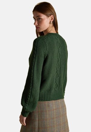Green knitted sweater featuring textured patterns, cable knit details on sleeves, ribbed hem, and a relaxed fit. Paired with a checkered skirt.