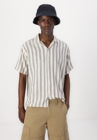 Kortærmet skjorte med knapper i lys beige med mørkeblå lodrette striber, sammen med khaki cargo shorts. Modellen bærer en sort bucket hat.