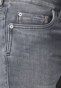 Grå denimjeans har en klassisk femficka design med synlig sömnad och metallnitar på fickorna. Texturerad tyg med en lätt blekning.