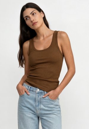 Woman with long dark hair wearing a sleeveless brown ribbed tank top and light blue jeans, hands in pockets, looking slightly to the side.