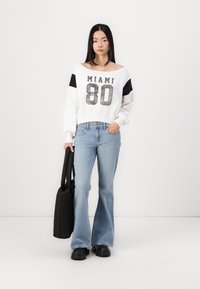 Sudadera blanca corta con gráfico "MIAMI 80", detalles en negro, jeans de campana azul claro y zapatos negros gruesos. Bolsa de mano negra.