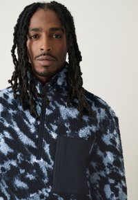 Fleece trui in blauw- en zwart tie-dye patroon, met een half-rits kraag en een effen zwart zakje op de borst.