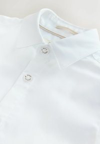 Balti marškiniai su apykakle, pagaminti iš tekstūruotos medžiagos; ant apykaklės yra du sidabriniai mygtukai su graviruotu prekės ženklu. Švelni apdaila ir paprastas dizainas.