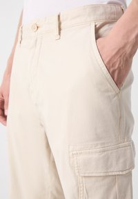 Pantalones cargo beige de algodón con bolsillos laterales, cierre de botón y detalle de costura visible. Textura suave, diseño casual.