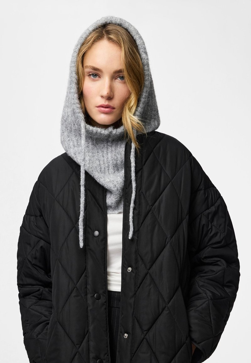 Joven con capucha de punto gris y chaqueta acolchada negra sobre una camiseta blanca, mirando hacia adelante con expresión neutral.