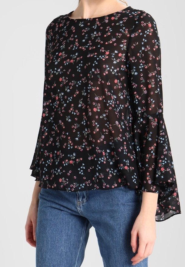 Femme portant un chemisier noir transparent avec de petits imprimés floraux rouges et bleus et des manches longues évasées, associé à un jean bleu.