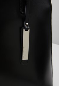 Sac à main en cuir noir à texture lisse, avec une étiquette en métal argenté attachée par une sangle noire, et des coutures précises le long des bords.