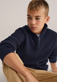 Mörkblå stickad tröja med dragkedja i kragen, med ett texturerat mönster och ribbade manschetter, bärs med beige byxor.