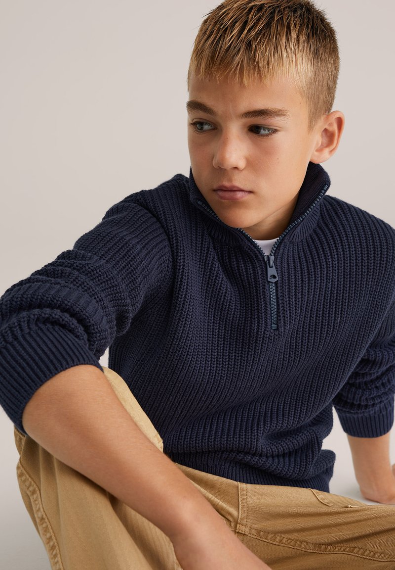 Mörkblå stickad tröja med dragkedja i kragen, med ett texturerat mönster och ribbade manschetter, bärs med beige byxor.