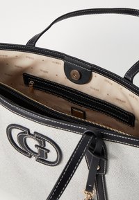 Sac à main en toile blanche avec des accents en cuir noir et un logo. Comprend une poche zippée intérieure et une doublure beige avec un motif "GUESS" répété.
