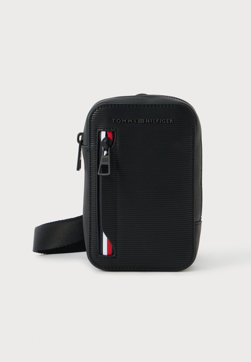 Bolso bandolera negro de nailon con textura acanalada, que presenta un bolsillo frontal con cremallera y un acento en rojo, blanco y azul. Correa ajustable.