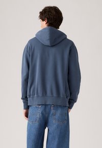 Mann som har på seg en blå hettegenser og blå denimjeans, stående med ryggen mot en ensfarget bakgrunn.