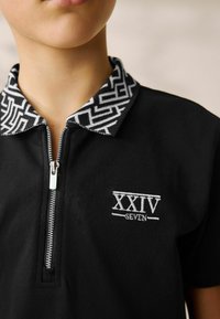 Polo nero con zip frontale, colletto a fantasia in bianco e nero e logo ricamato "XXIV SEVEN" in bianco.
