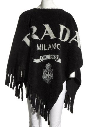 Czarny dzianinowy ponczo z frędzlami na brzegach, biały napis "PRADA MILANO DAL 1913" i herb na przodzie, noszone na białych spodniach.