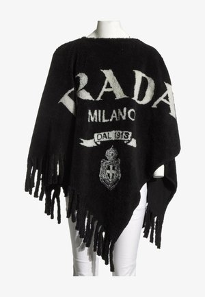 Zwart gebreide poncho met franjes aan de randen, witte tekst "PRADA MILANO DAL 1913" en wapenlogo aan de voorkant, gedragen over een witte broek.