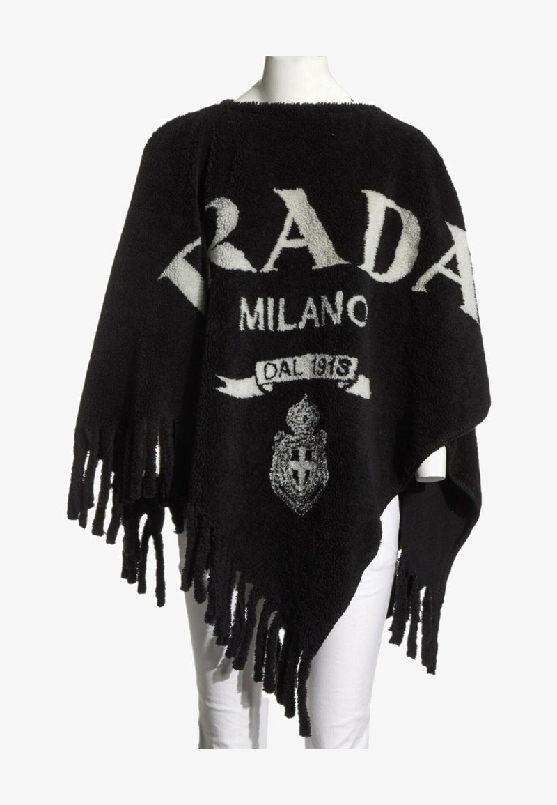 Poncho en tricot noir avec bords à franges, texte blanc « PRADA MILANO DAL 1913 » et logo écusson sur le devant, porté par-dessus un pantalon blanc.