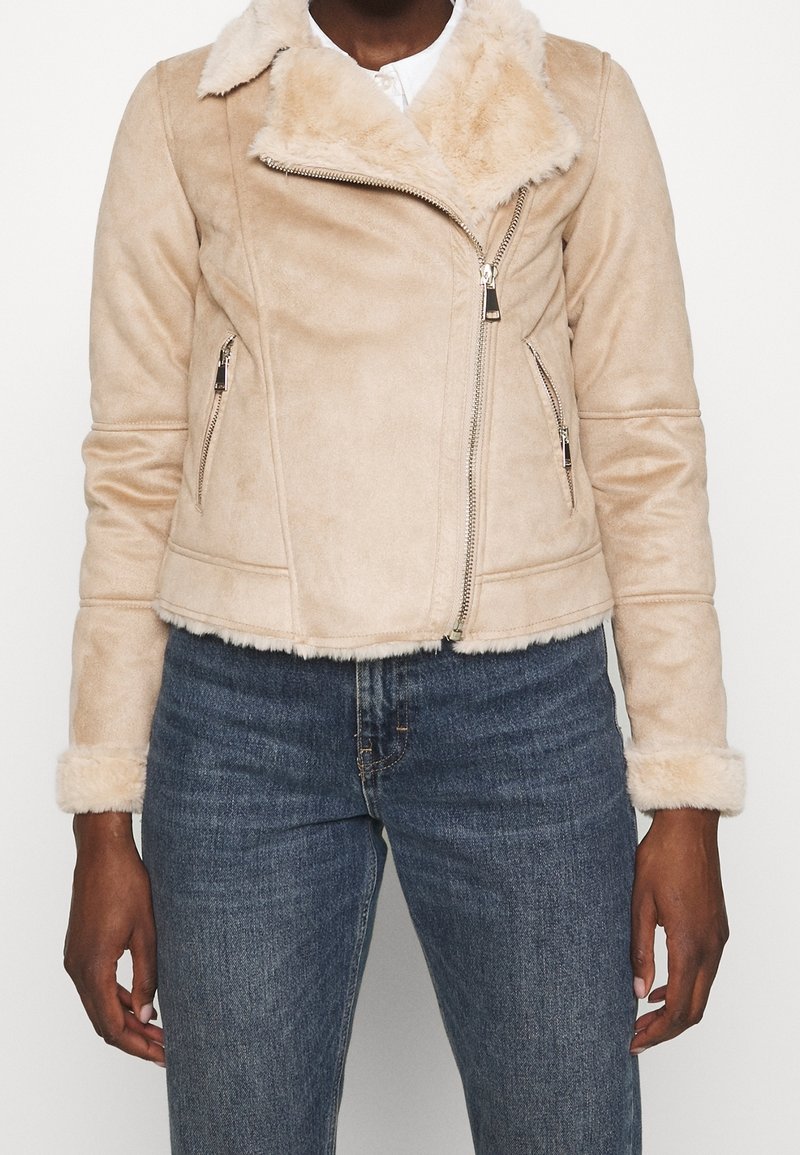 Chaqueta de piel sintética beige con cuello de muesca, cierre de cremallera y bolsillos laterales; presenta puños y ribetes de pelaje. Textura suave y acogedora.