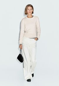 Helles beiges Sweatshirt mit Rundhalsausschnitt, kombiniert mit weiten weißen Hosen. Das Model hält eine kleine schwarze Handtasche und trägt schwarze Schuhe. Minimalistisches Design.