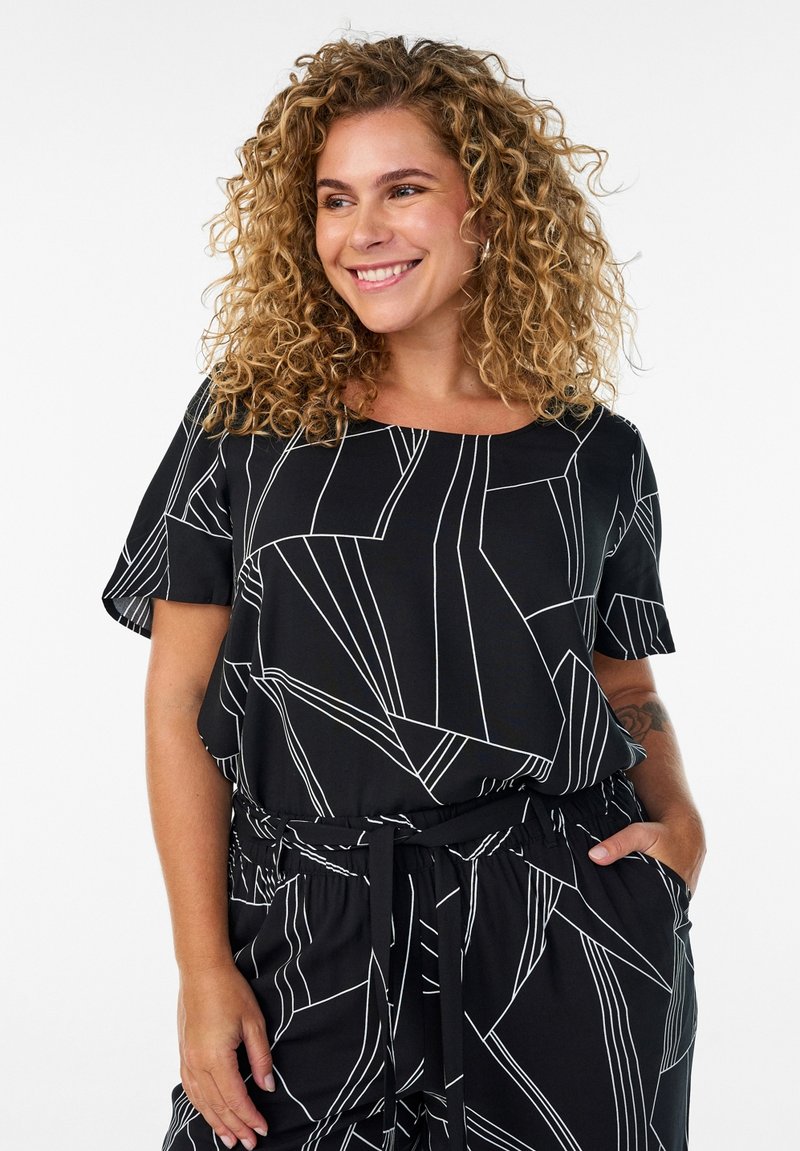 Donna sorridente con capelli ricci, indossa un top nero e pantaloni con motivi a linee geometriche bianche, con una mano in tasca, su sfondo bianco.