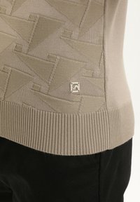 Beige tekstuuriga kudum, millel on geomeetriline kolmnurga muster ja soonikkoesäär, kantud mustade pükste peal.