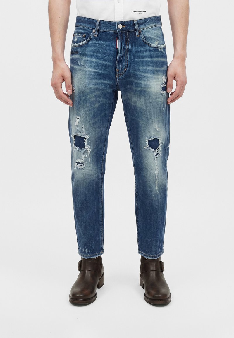 Vue de face d'un jean bleu usé avec des déchirures et des patchs, associé à des bottines marron foncé sur une personne.