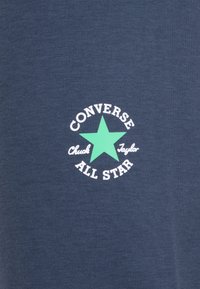 Navy blue puuvillapaita, jossa on valkoinen pyöreä logo vihreällä tähdellä, ja tekstinä "CONVERSE Chuck Taylor ALL STAR" tähden alapuolella.