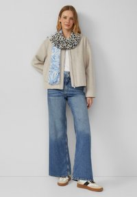 Veste en laine beige zippée, écharpe florale bleue, jeans bleu clair à jambe large, et baskets multicolores avec une semelle texturée.