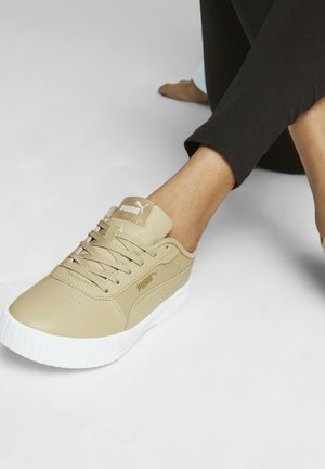 Person trägt beige Puma-Sneaker mit weißen Sohlen und schwarzen Hosen, sitzt mit kreuzbeinigen Beinen auf einem schlichten weißen Boden.