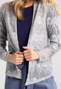 Spetsjacka i ljusgrått med blommönster, lättviktsmaterial, öppen framdesign och långa ärmar över en mörk topp.