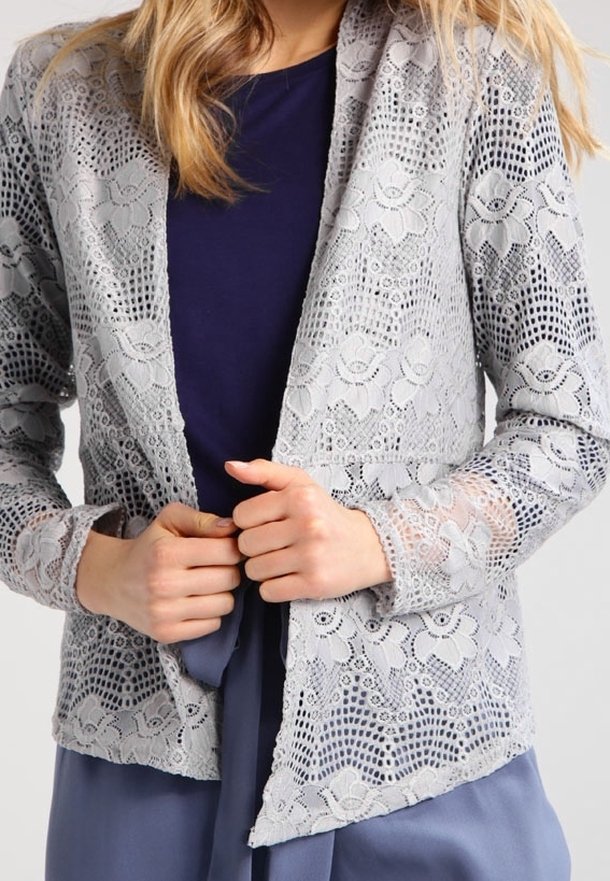 Spetsjacka i ljusgrått med blommönster, lättviktsmaterial, öppen framdesign och långa ärmar över en mörk topp.