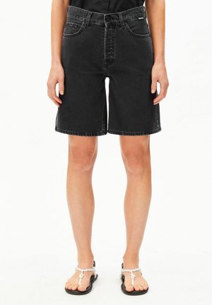 Shorts en denim noir longueur genou portés par une personne debout, assortis à des sandales blanches à lanières en corde et un haut noir.