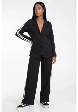 Kobieta w czarnym garniturze z białymi paskami na rękawach i spodniach, w połączeniu z czarno-białymi sneakersami, stoi z jedną ręką na biodrze.
