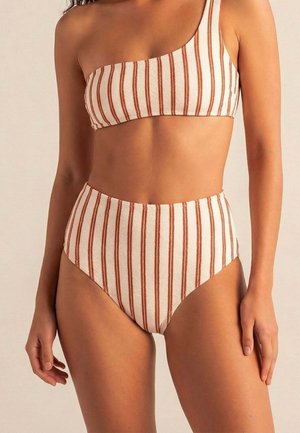 Mujer con un bikini de talle alto y un solo tirante, con rayas verticales marrones y blancas, de pie frente a un fondo liso.