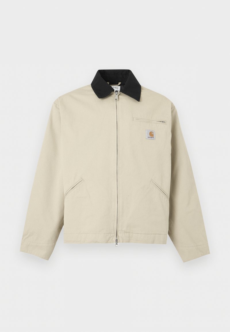 Chaqueta Carhartt beige con cuello negro, cremallera completa en el frente, dos bolsillos laterales y un pequeño bolsillo con cremallera en el pecho con parche de logo.
