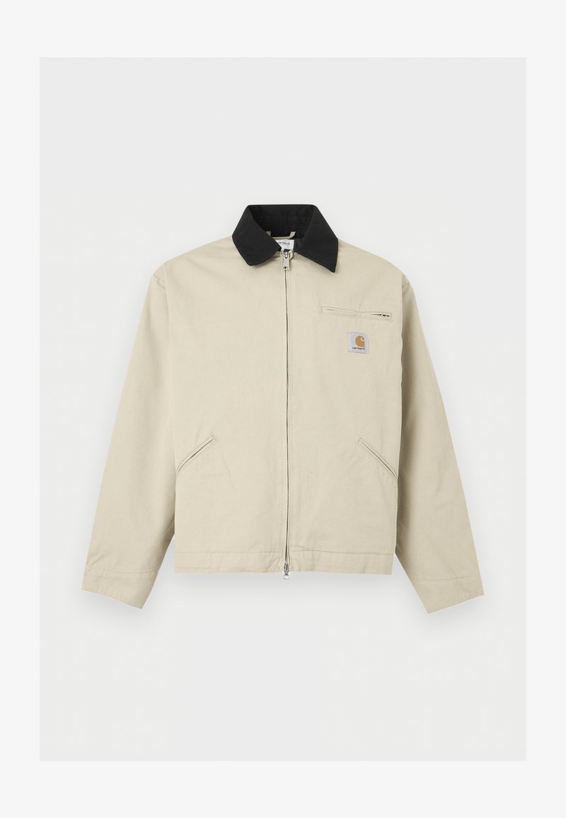 Chaqueta Carhartt beige con cuello negro, cremallera completa en el frente, dos bolsillos laterales y un pequeño bolsillo con cremallera en el pecho con parche de logo.