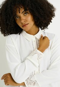 Blusa blanca con un acabado texturizado, que presenta un cuello y puños con bordado de encaje, una tapa de botones vertical y detalles de ojales decorativos.